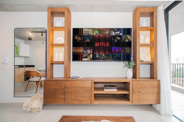 A modern living room with a wooden entertainment unit, a flat-screen TV mounted above it, and a view of the kitchen in the background. - Stylish City Escape with Panoramic Views, Pool&Gym (Asunción)