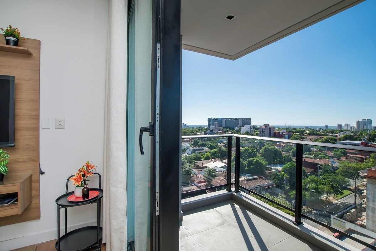 Een balkon met uitzicht op de stad, een tafeltje met een bloevas en een wandtelevisie.
