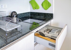 Une cuisine avec un plan de travail noir, une bouilloire en acier inoxydable, un égouttoir et un tiroir contenant des ustensiles de cuisine.