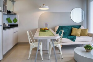 Un salon moderne avec un canapé blanc, une table à manger avec des chaises, une petite table ronde agrémentée d’une plante, et un coin cuisine avec des meubles blancs et un luminaire suspendu.