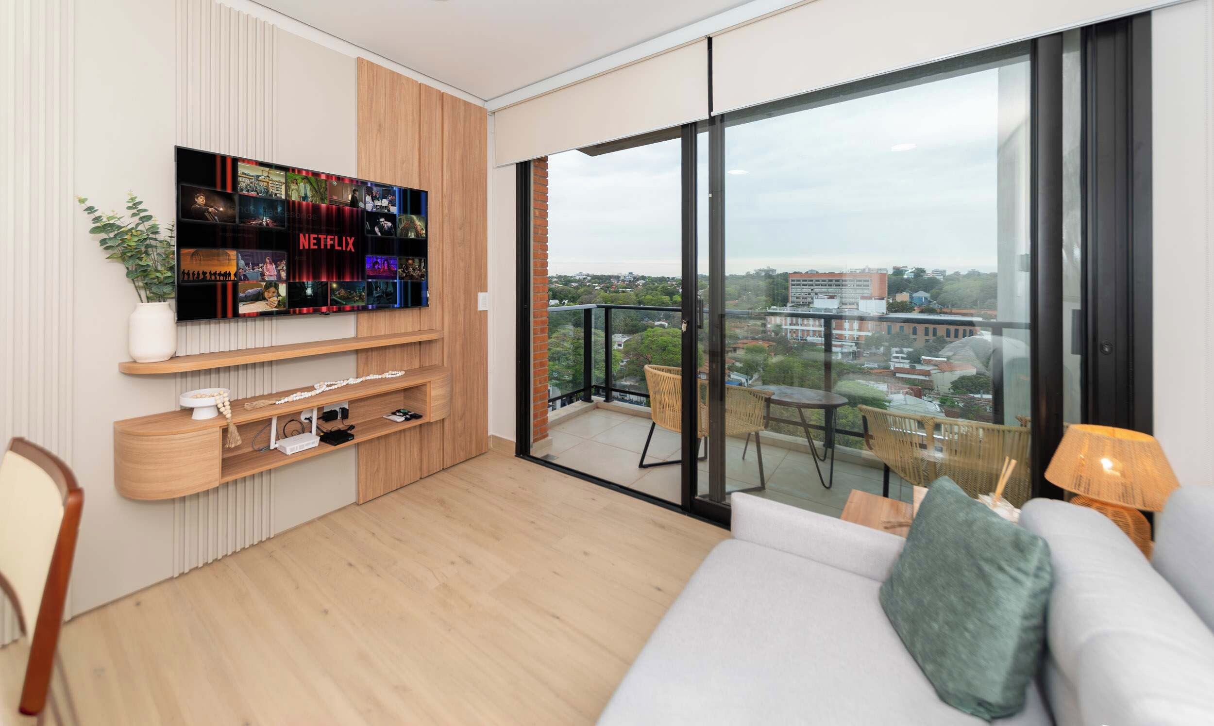 Una sala moderna con un televisor grande montado en la pared, un sofá, un comedor con mesa y sillas, y un balcón con vista a la ciudad.