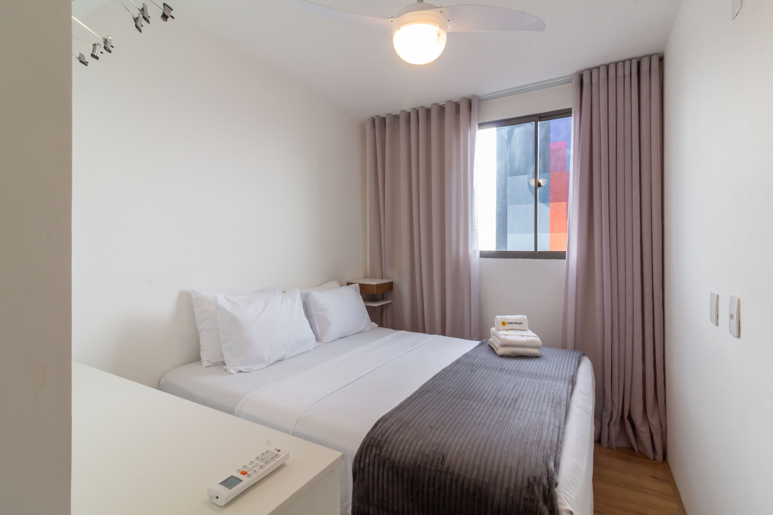 Une chambre d’hôtel avec un lit, une table de chevet, une télécommande, une fenêtre avec des rideaux et un ventilateur de plafond.