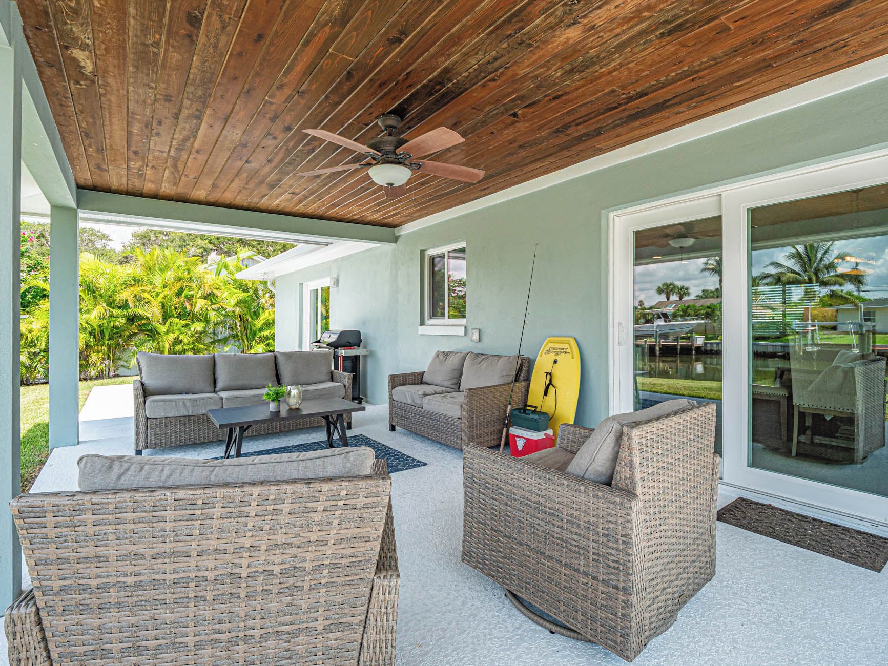 Une terrasse extérieure couverte, aménagée avec des meubles en osier, un ventilateur de plafond et offrant une vue sur une piscine et un espace verdoyant.
