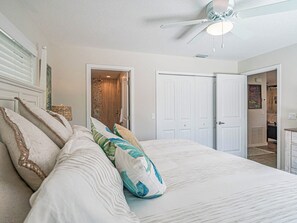 A bedroom with a large bed, white walls, a ceiling fan, and a door leading to another room. - Waterfront 3BR home with private dock, down the street from the beach! (Fort Pierce)