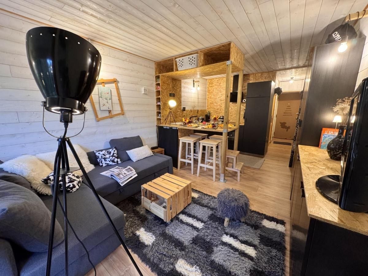 A cozy living room with a sofa, coffee table, and a lamp. There is a kitchen area with stools and a dining table.