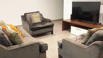 Una sala de estar con dos sofás grises, un sillón pequeño con un cojín y un televisor de pantalla plana sobre un mueble de madera.