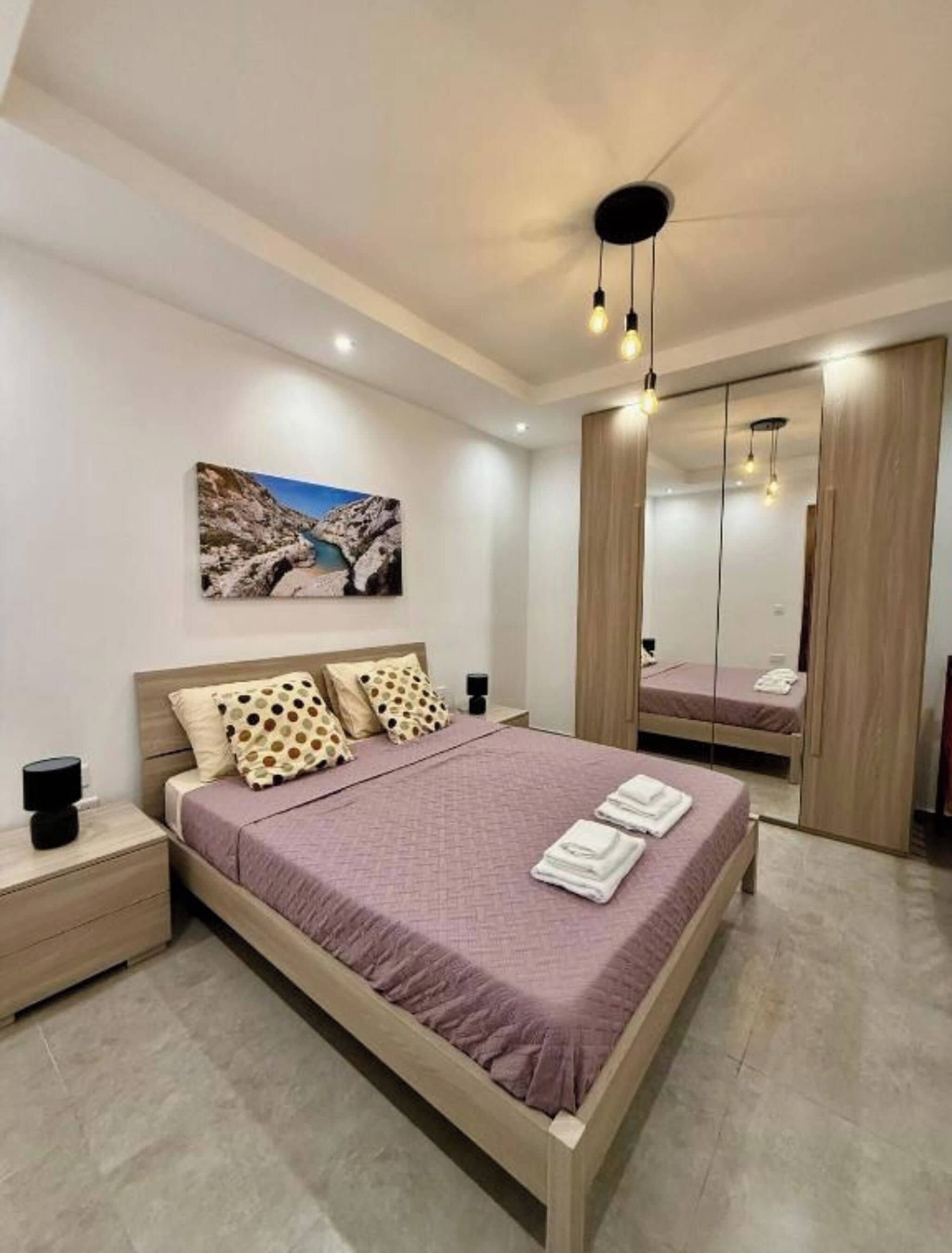 A modern bedroom with a large bed, a nightstand, a bedside table, a mirror, and a painting on the wall.