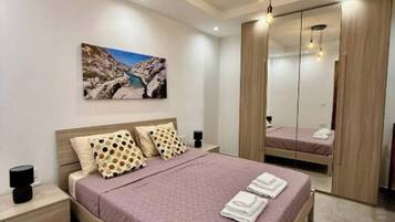 A modern bedroom with a large bed, a nightstand, a bedside table, a mirror, and a painting on the wall.
