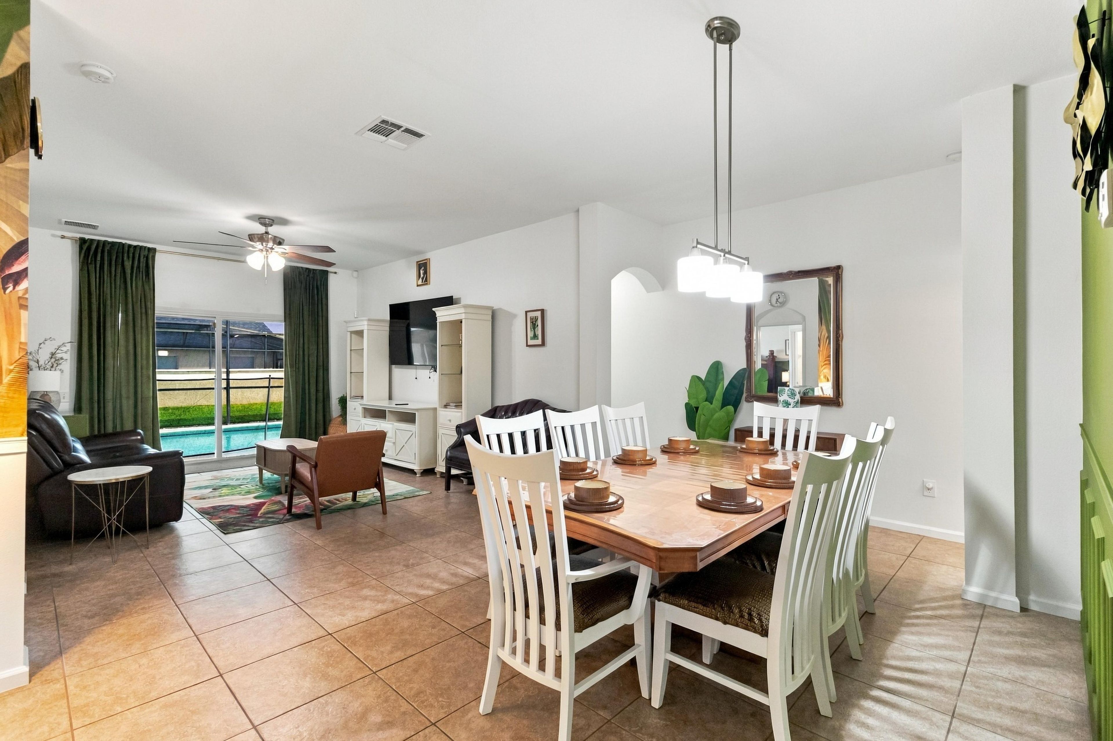 Un vaste espace dédié à la salle à manger, avec une grande table en bois et des chaises blanches, un ventilateur de plafond et une vue sur une piscine.