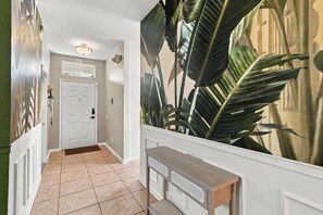 A hallway with a door, a wall-mounted light fixture, a console table, and a large plant. - 4/2 Vacation Home at Windsor Palms!! (Kissimmee)