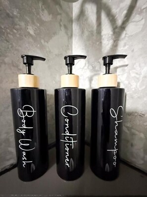Tres botellas negras con dosificadores tipo bomba, etiquetadas 'Body Wash', 'Conditioner' y 'Shampoo', frente a una pared con textura.