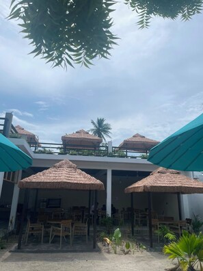 Un área de estar al aire libre con sombrillas de paja y sillas de madera, una piscina al fondo y un cielo despejado.