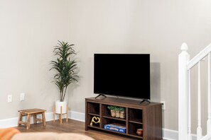 Ein Wohnzimmer mit einem Flachbildfernseher auf einem Holzständer, einem kleinen Holztisch, einer Topfpflanze und einem Spielkarton auf dem Ständer.