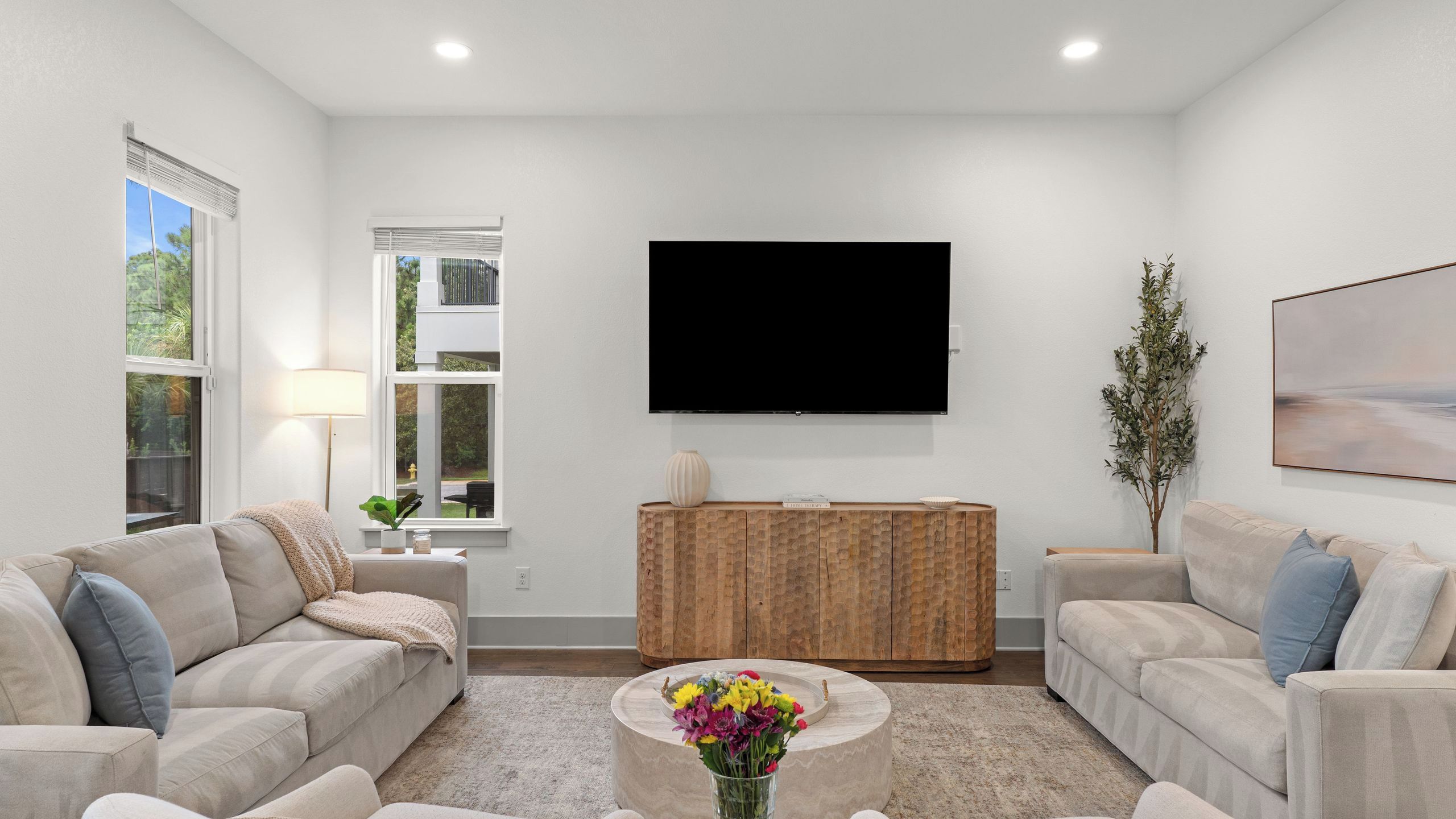 A modern living room with two sofas, a coffee table with flowers, a TV, and a window with blinds.
