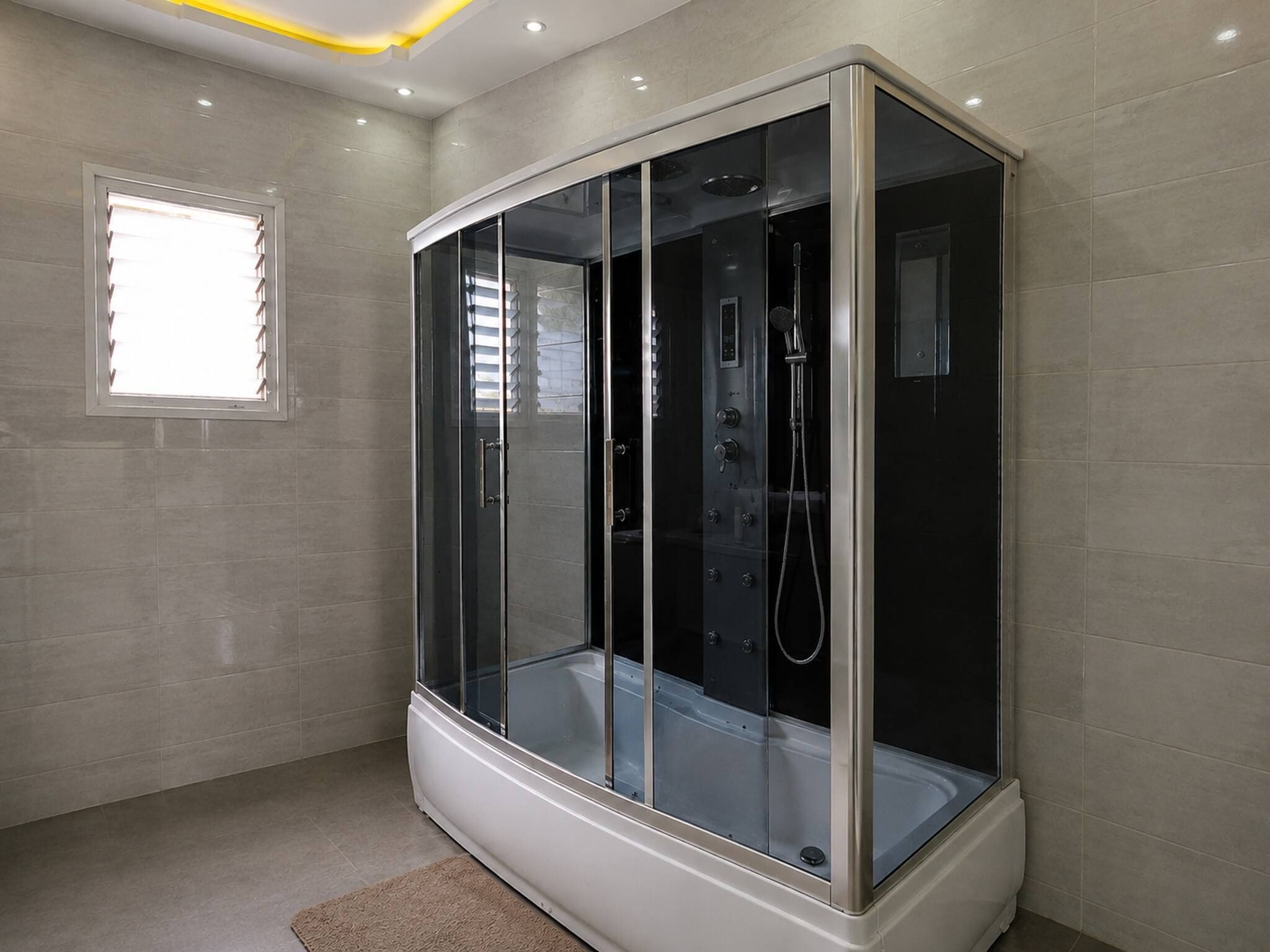 Un baño moderno con ducha envuelta en vidrio, una ventana con persianas y piso de tonos claros.