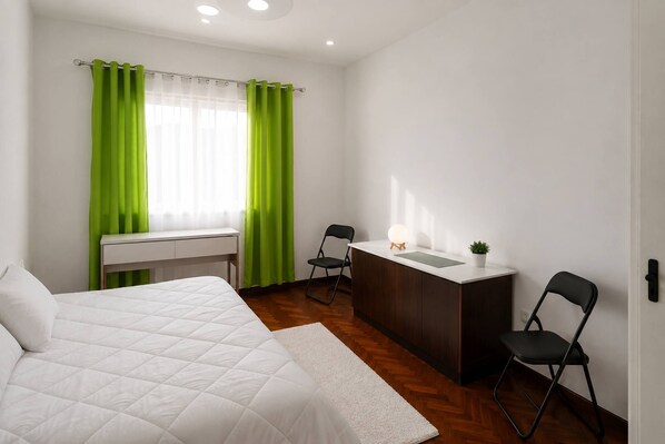 A bedroom with a bed, a desk, a chair, a small table with a lamp, and a potted plant.