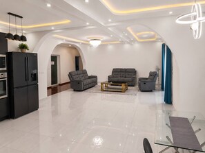 A modern living room with a glass dining table, two grey sofas, a kitchen area with a black refrigerator, and a blue curtain.