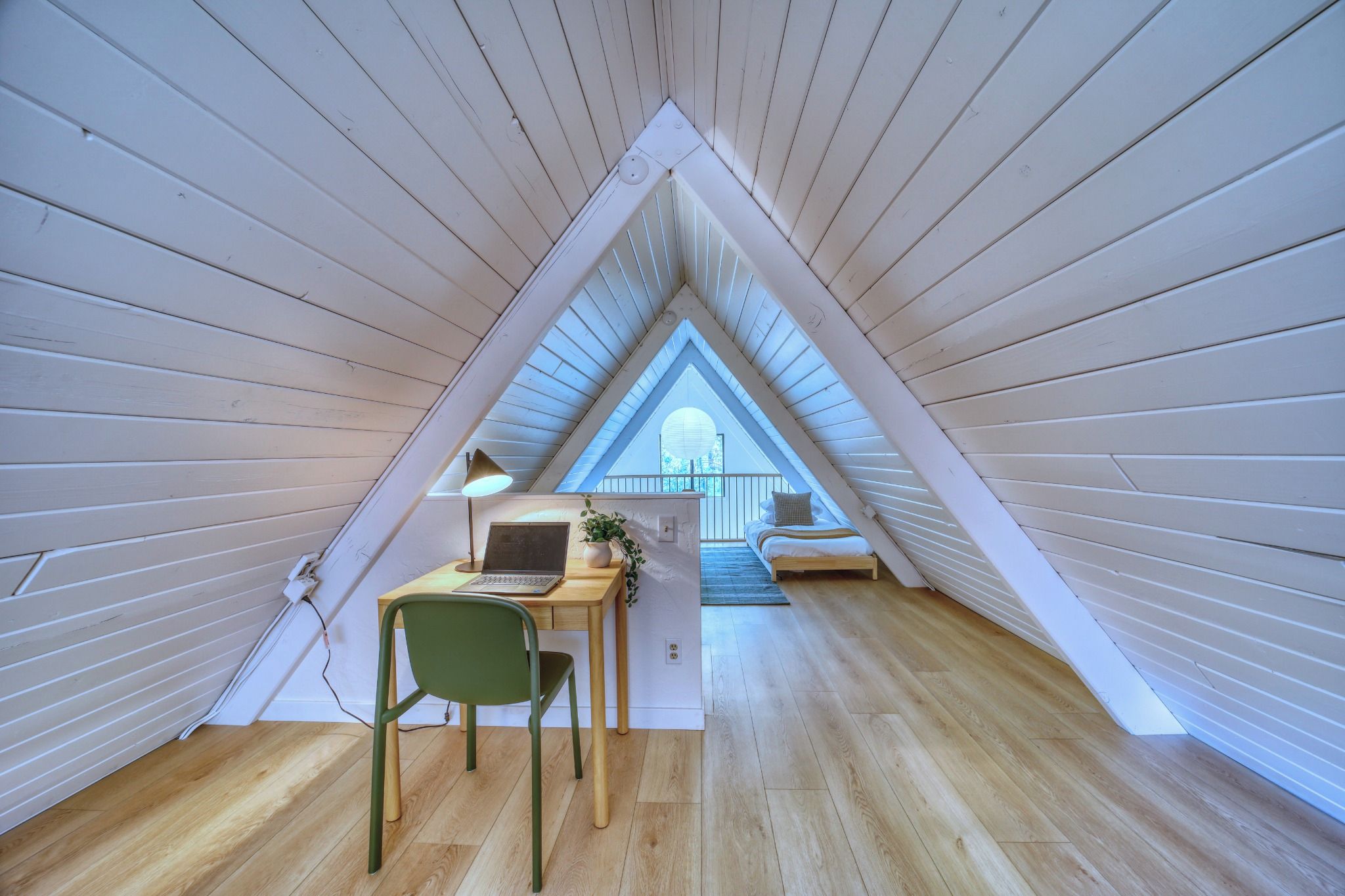 Ein quadratisches Schlafzimmer im Dachgeschoss mit Holzboden, einem Schreibtisch mit Laptop, einem Stuhl und einem Bett mit Blick zum Himmel.