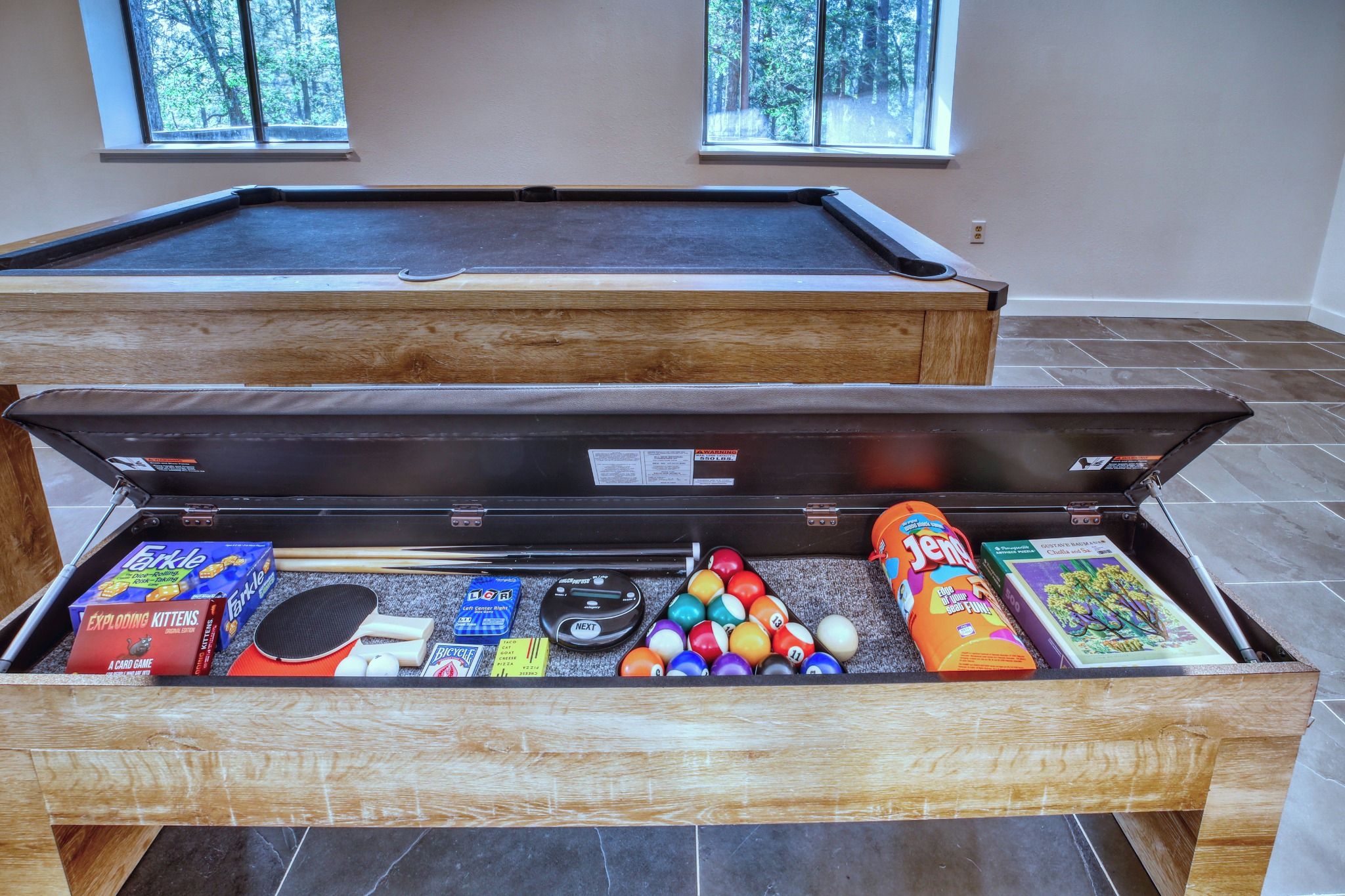 A pool table with a wooden side table in front containing various pool balls, a paddle, and a card game.