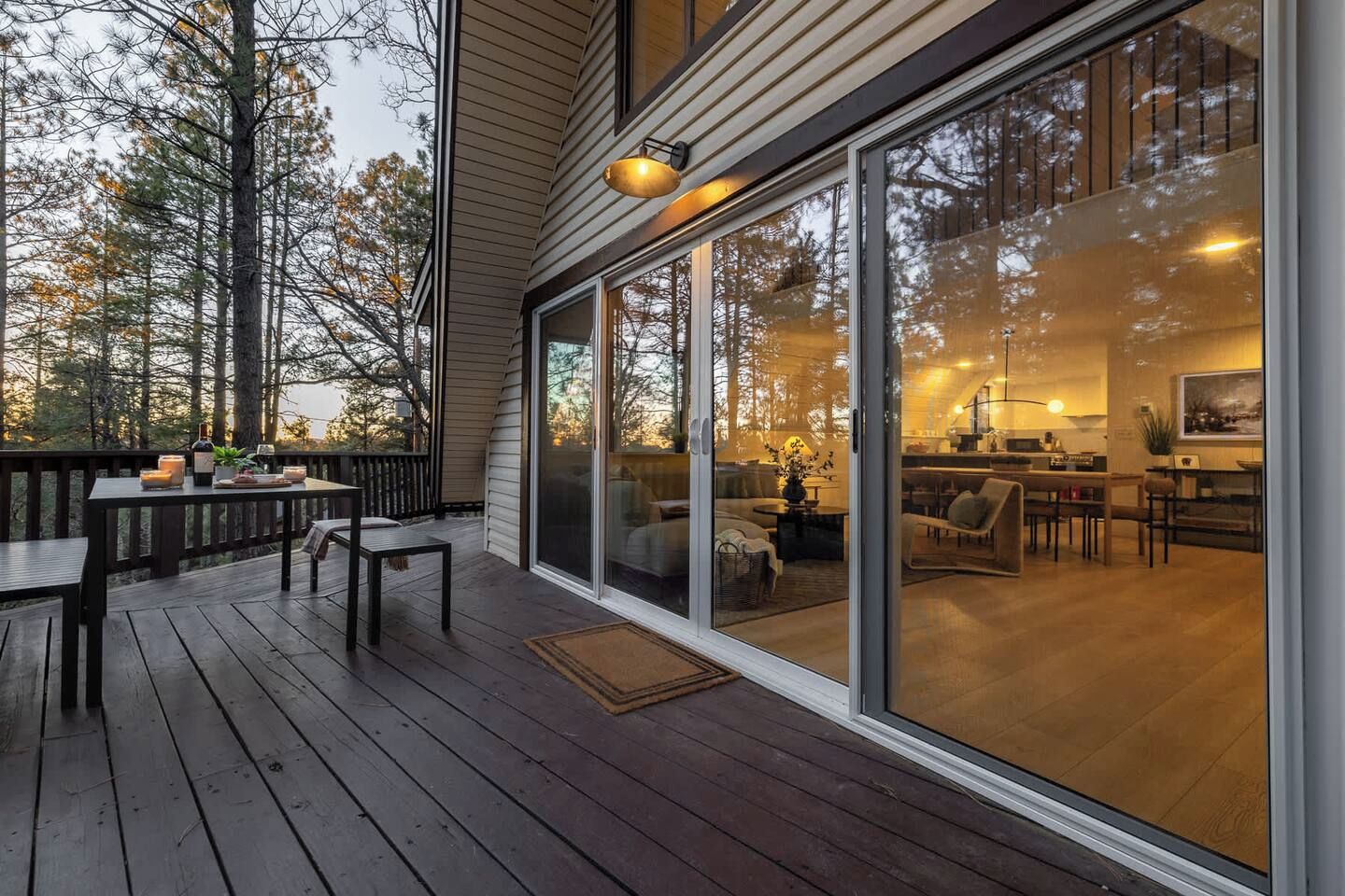 Une terrasse en bois avec une table et des chaises, une porte-fenêtre coulissante donnant sur un espace de vie avec un canapé et une table basse, et une cuisine visible à travers la vitre.