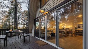 Eine Holzdachterrasse mit Esstisch und Stühlen, eine Glastür, die zu einem Wohnbereich mit Sofa und Couchtisch führt, und eine durch die Scheibe sichtbare Küche.