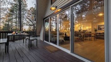 Une terrasse en bois avec une table et des chaises, une porte-fenêtre coulissante donnant sur un espace de vie avec un canapé et une table basse, et une cuisine visible à travers la vitre.
