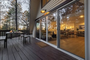 A wooden deck with a dining table and chairs, a sliding glass door leading to a living area with a sofa and a coffee table, and a kitchen visible through the glass.