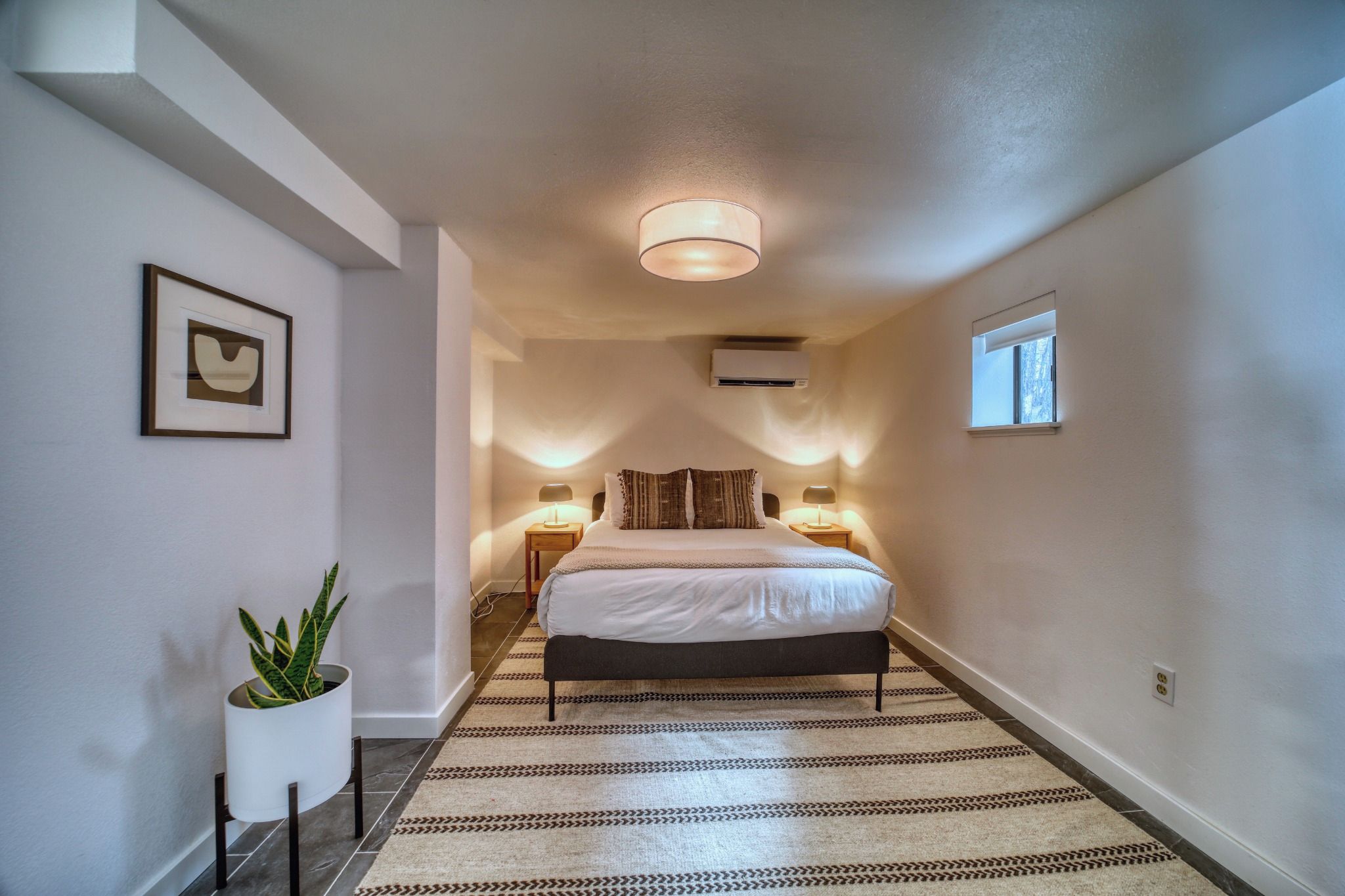 A hotel room with a bed, bedside lamps, a potted plant, and a framed picture on the wall.