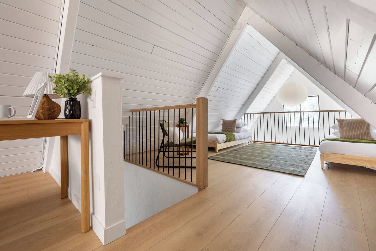 Une chambre à coucher aménagée dans un grenier, avec un plafond en pente, un plancher en bois, un lit, un coin salon et une petite table agrémentée d’une plante.
