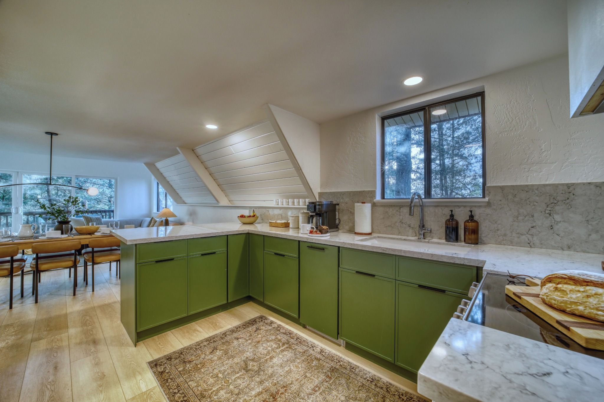 A modern kitchen with green cabinets, a marble countertop, and a large window overlooking a forest.