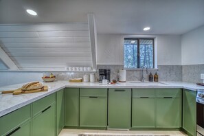 Una cocina con armarios verdes, encimera de mármol, cafetera y ventana con vistas a árboles.