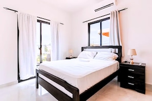 Quarto de hotel com cama, mesa de cabeceira, abajur e janela com cortinas.