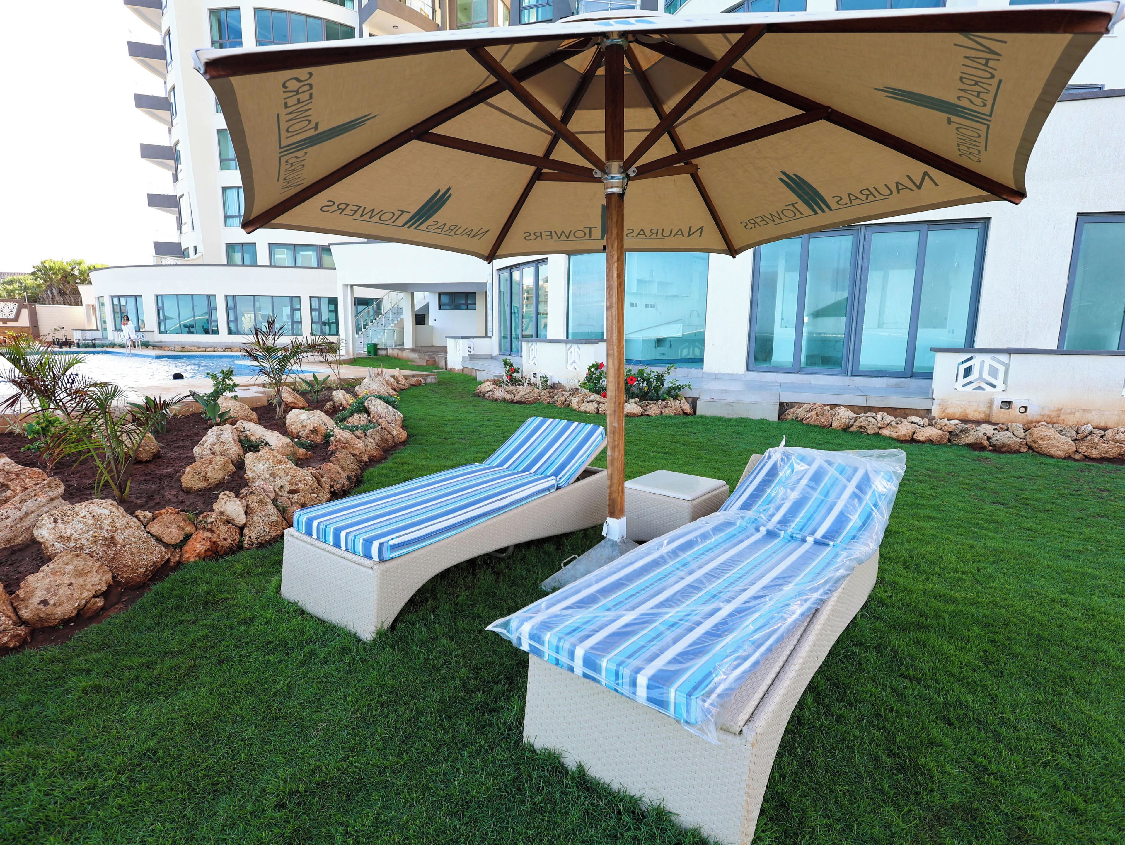 A poolside area with lounge chairs and a large umbrella, surrounded by a well-maintained lawn and a modern building in the background.