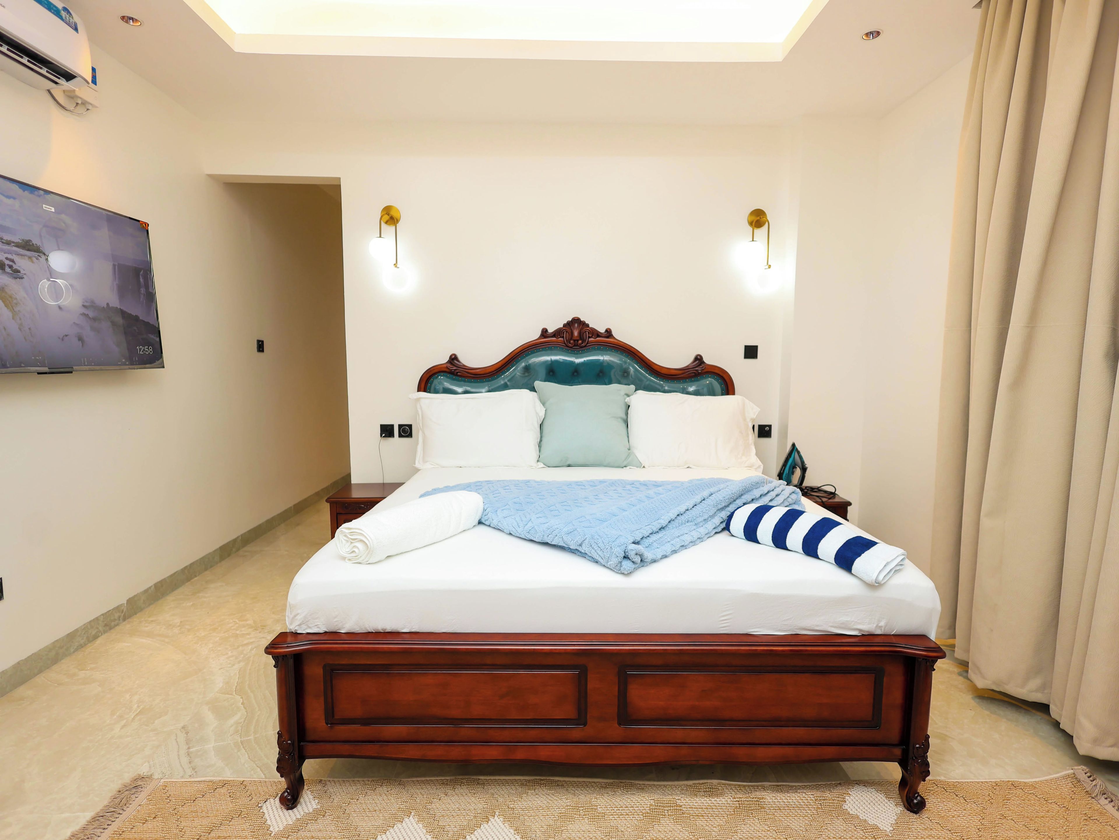A neatly made bed with a wooden headboard, a wall-mounted flat-screen TV, and two wall sconces.