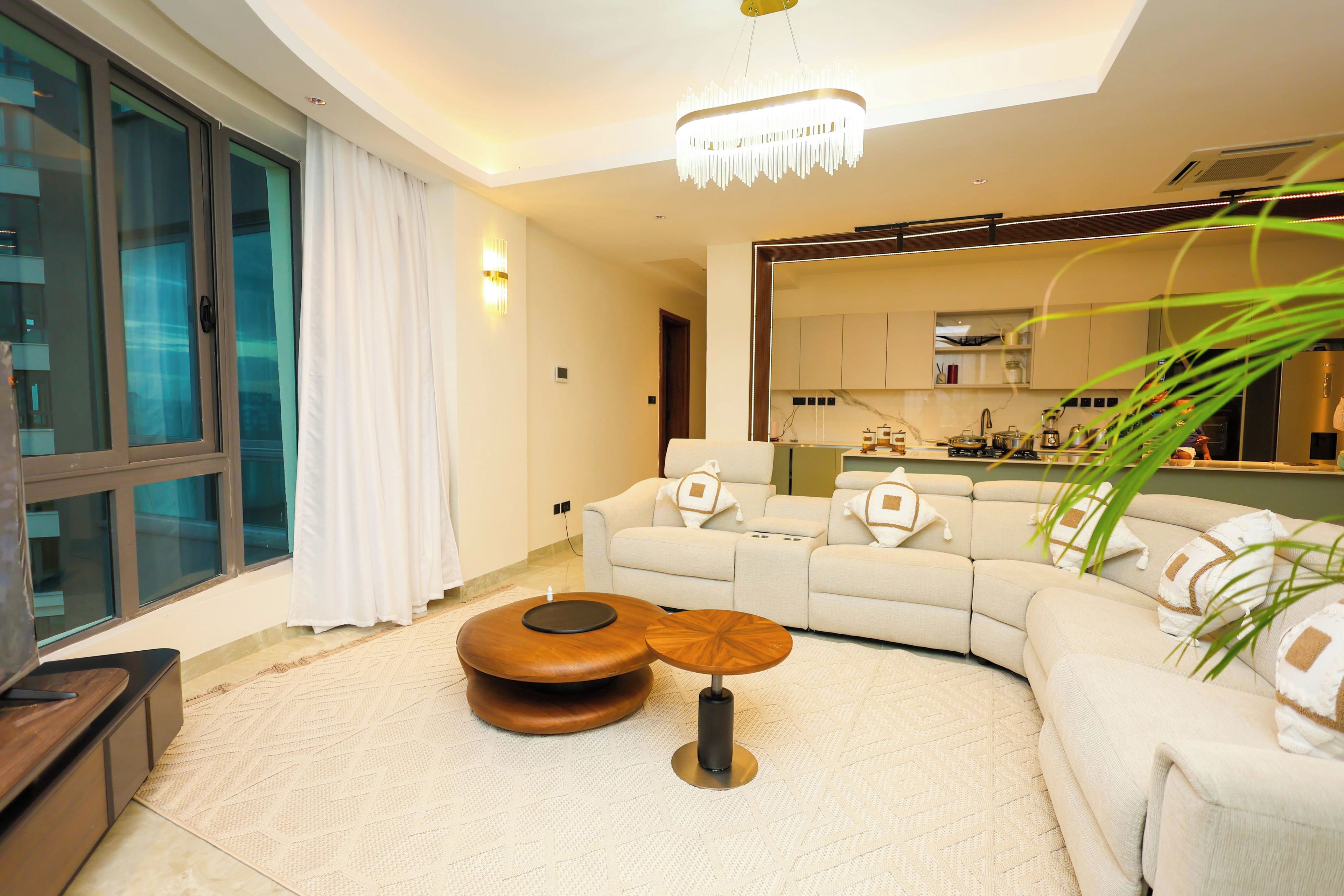 A modern living room with a large window, a sofa, a coffee table, and a kitchen area visible through an open door.