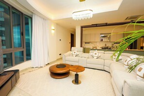 A modern living room with a large window, a sofa, a coffee table, and a kitchen area visible through an open door.