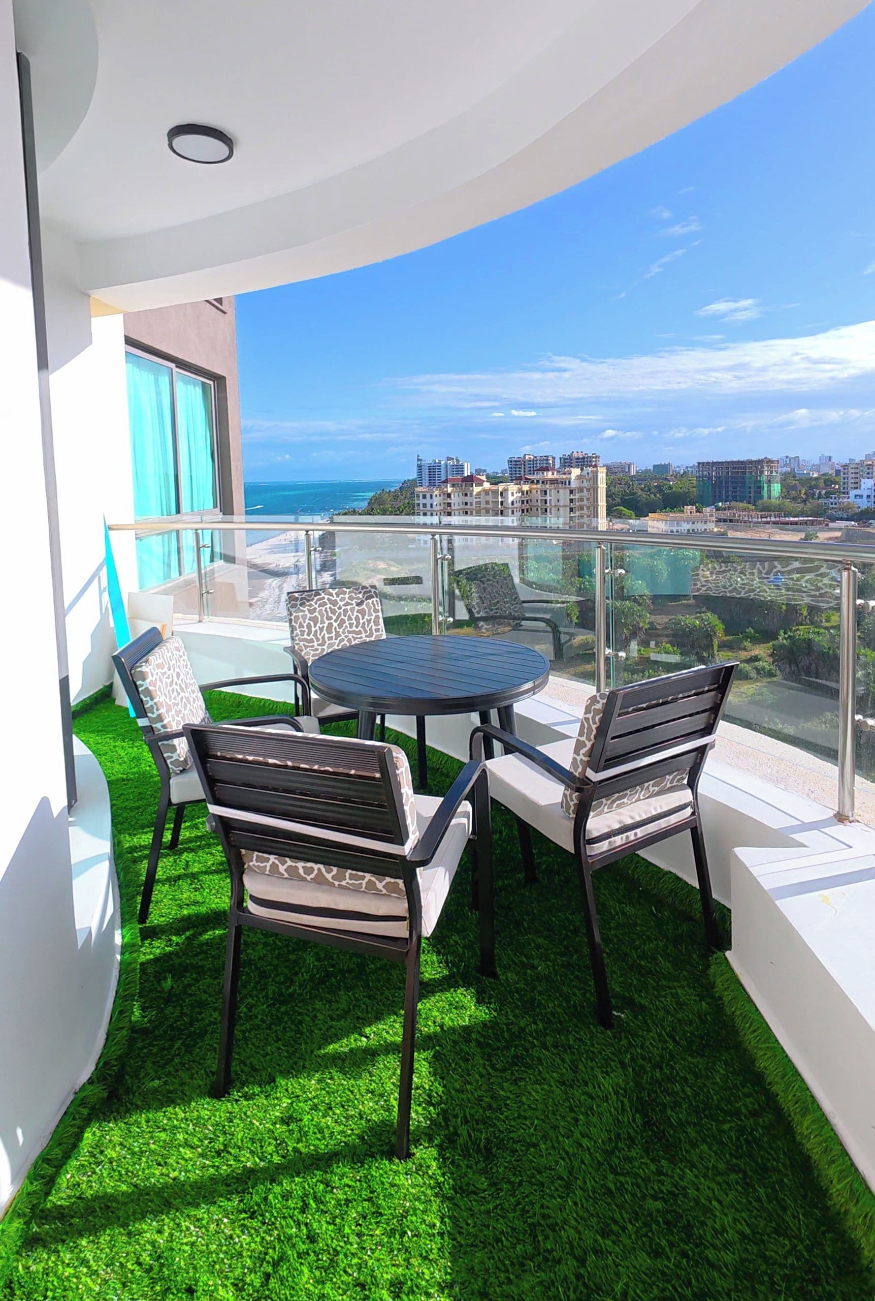 A balcony with a glass railing, a round table, and four chairs with patterned cushions. The balcony has artificial green grass and overlooks a cityscape with buildings and a body of water.