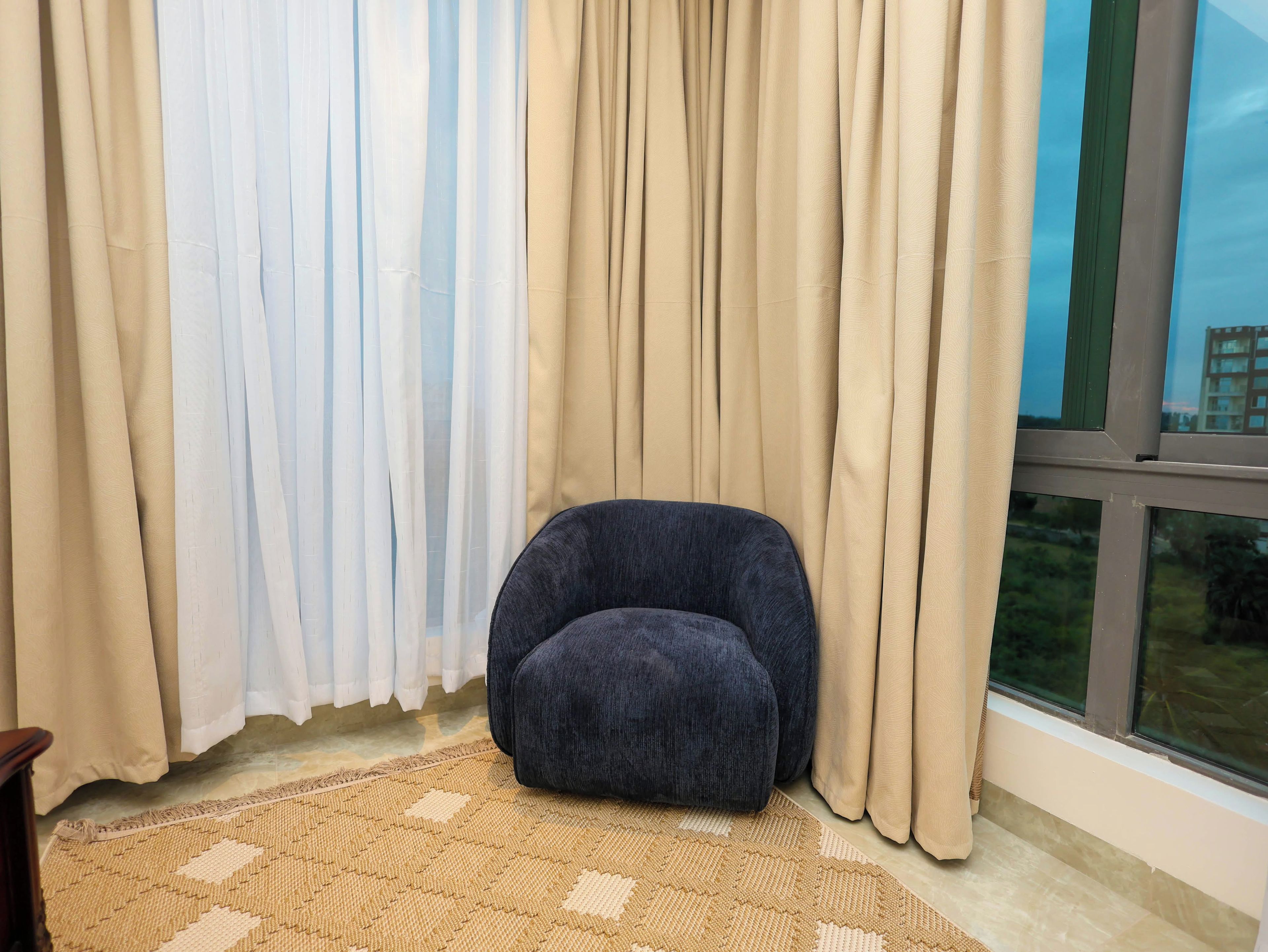 Una habitación con una silla azul oscuro, cortinas color beige y vistas por una ventana.