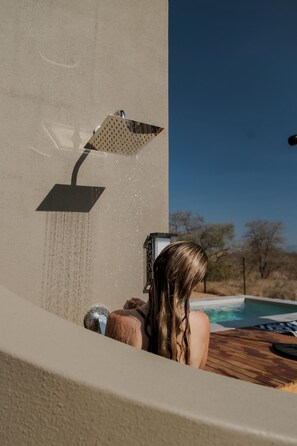 A person in a bathtub with a showerhead above, a pool in the background, and a clear sky.