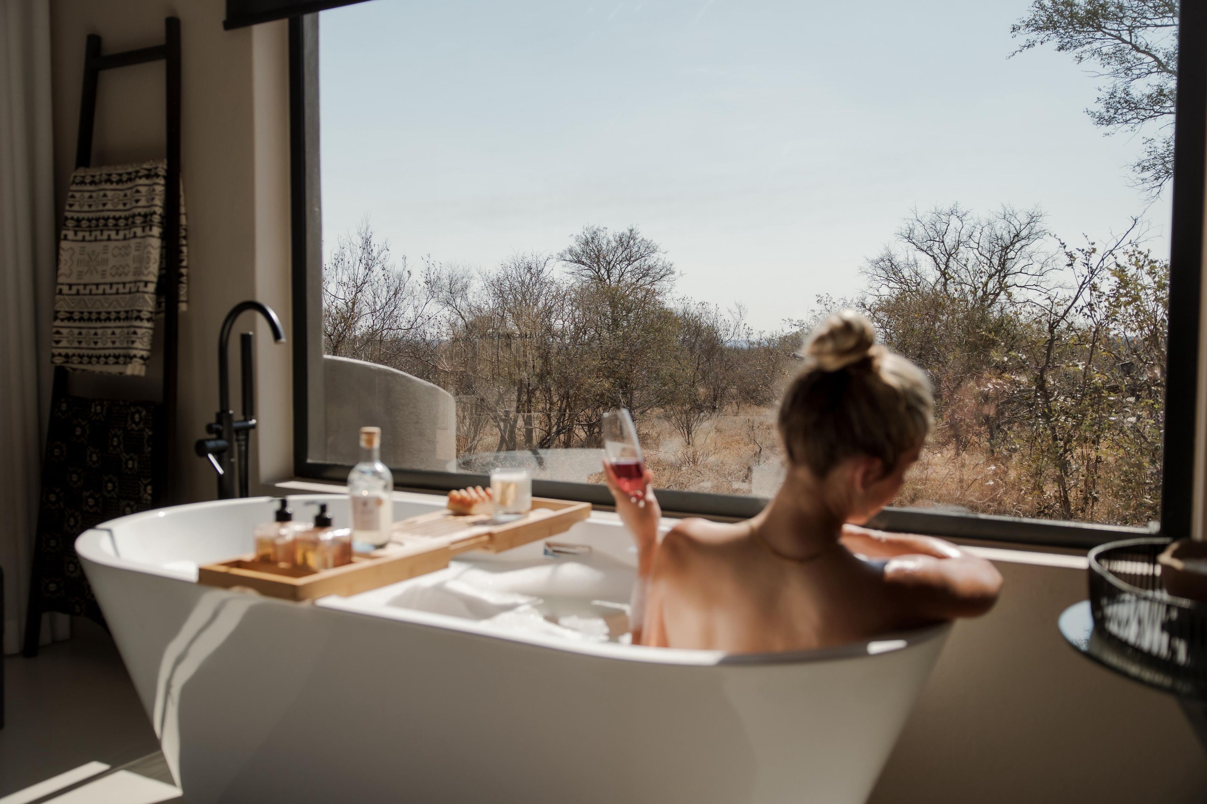 Une personne se détend dans une baignoire, avec une tablette contenant des produits de toilette, une bouteille et un verre de vin rouge, et peut admirer les arbres à travers la fenêtre.