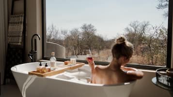 Une personne se détend dans une baignoire, avec une tablette contenant des produits de toilette, une bouteille et un verre de vin rouge, et peut admirer les arbres à travers la fenêtre.