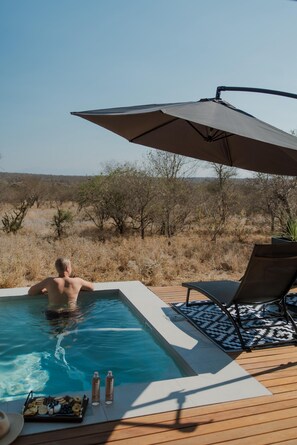 A man in a swimming pool with a wooden deck, lounge chairs, and an umbrella.