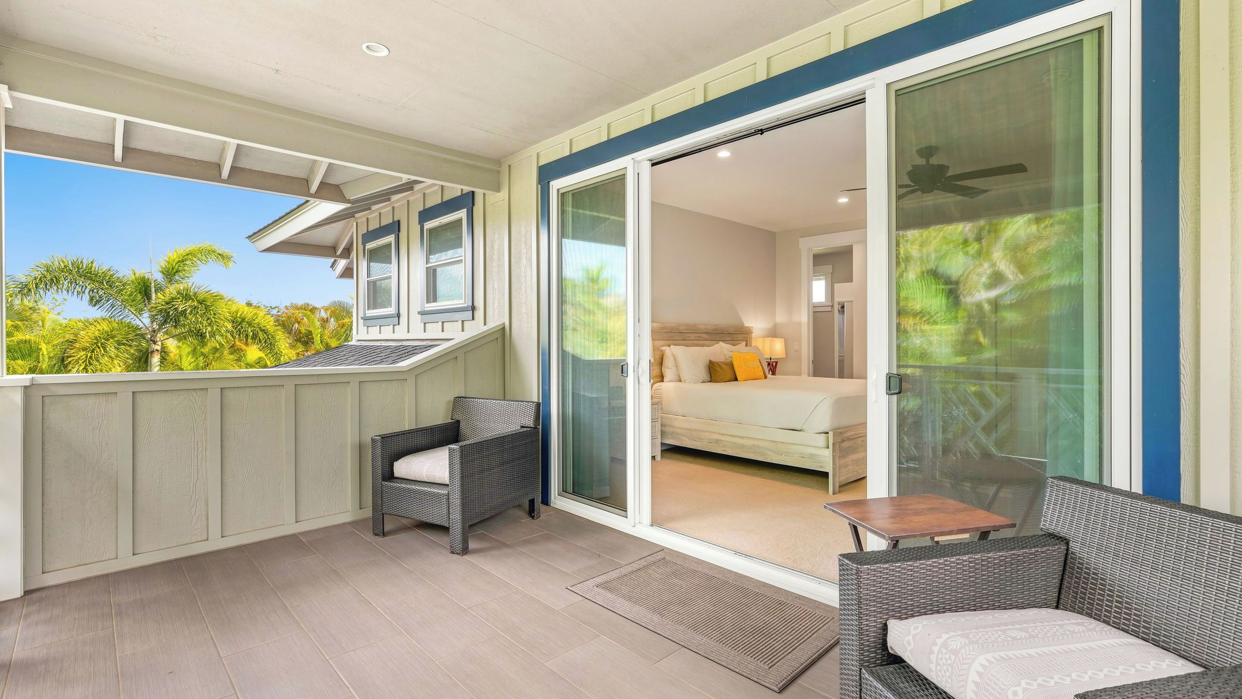A balcony with wicker furniture, a sliding glass door leading to a bedroom with a bed and a nightstand, and a view of greenery outside.