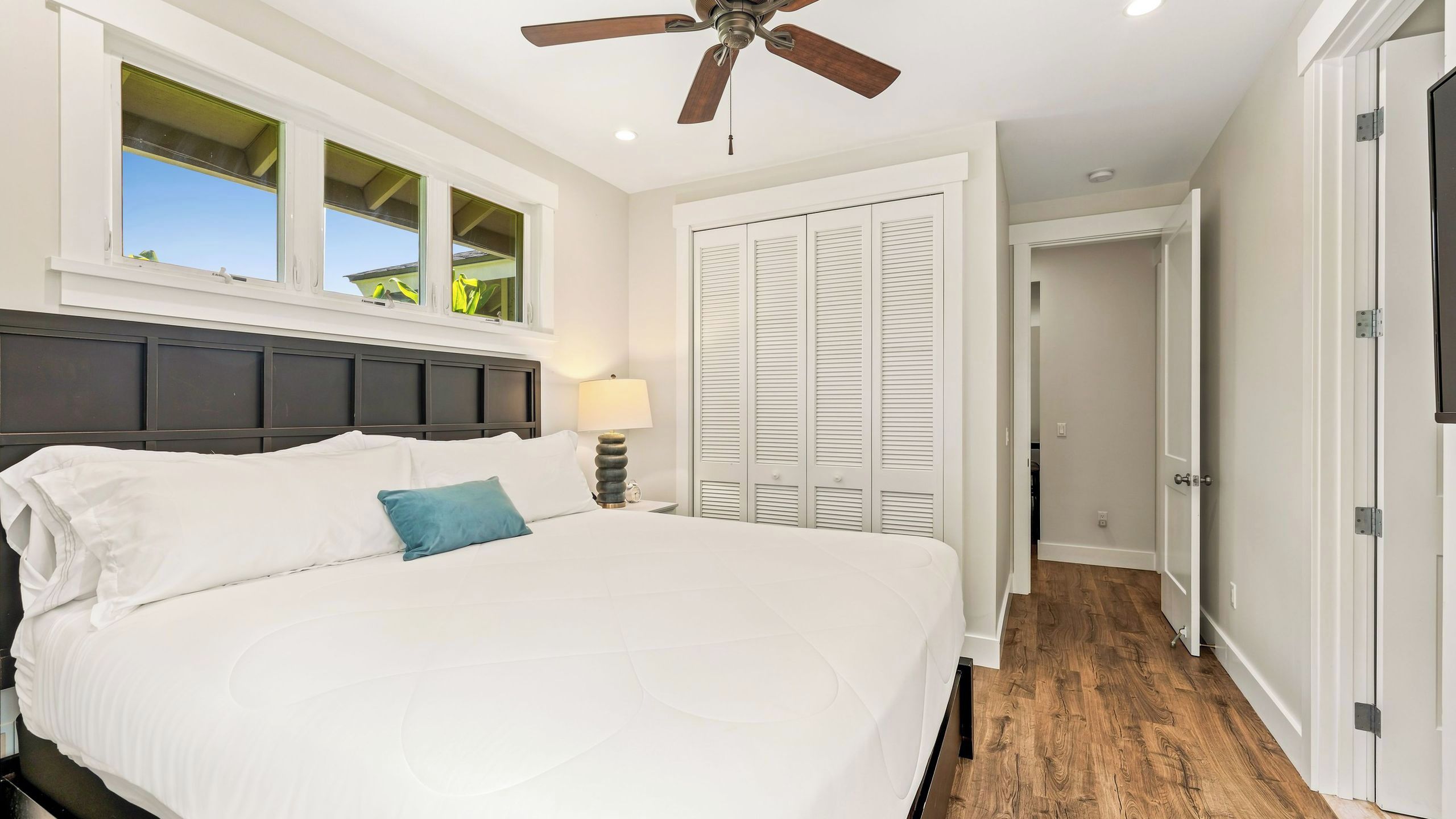 A bedroom with a bed, a ceiling fan, a window with blinds, and a door leading to another room.