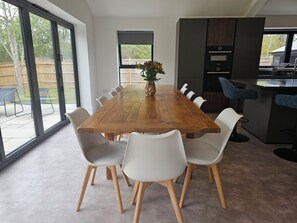 Un comedor con una mesa de madera y sillas blancas, un jarrón con flores y vistas al exterior a través de amplias ventanas.