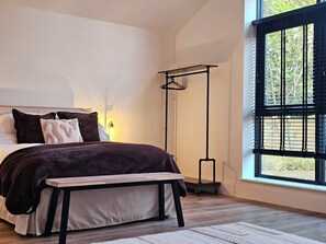 Un dormitorio con cama, mesita de noche, banco, lámpara de pie y ventana con persianas.