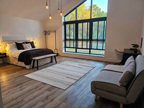 A modern bedroom with a large window, a bed, a bench, a sofa, and a desk.