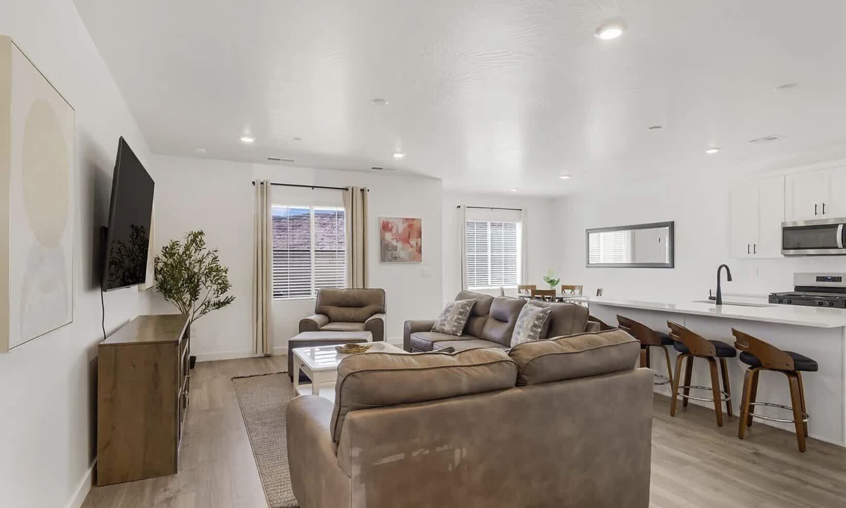A modern living room with a sofa, coffee table, and TV. There is a kitchen area with a dining table and bar stools.