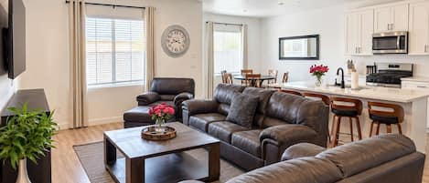 A modern living room with leather sofas, a coffee table, and a TV. There is a kitchen area with white cabinets and a microwave.