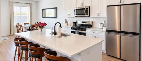 A modern kitchen with white cabinetry, stainless steel appliances, a central island with bar stools, and a dining area with a table and chairs.